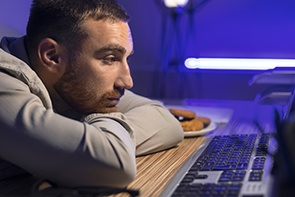 Un homme est assis devant un ordinateur, appuyé sur son bureau, le visage fatigué. Il regarde fixement l’écran avec un air pensif ou épuisé. La scène est éclairée par une lumière bleue violacée. On distingue un clavier d’ordinateur au premier plan, et une assiette contenant quelques biscuits ou cookies à côté de lui. L’homme a une barbe courte et des cheveux bruns coupés courts, il porte une chemise claire.