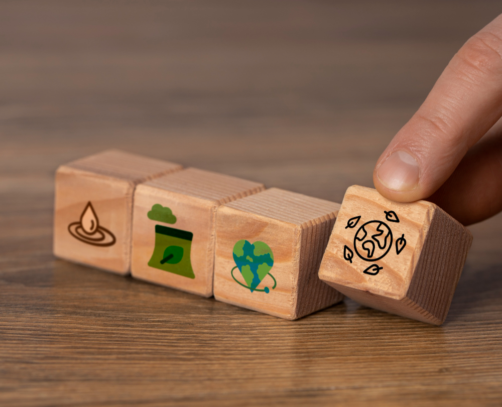 Image montrant quatre cubes en bois alignés sur une table.
Chaque cube porte une icône liée à l’environnement : une goutte d’eau, un sac ou contenant recyclable, un cœur représentant la planète, et un globe entouré de feuilles.
Une main saisit le dernier cube, celui avec le globe.
