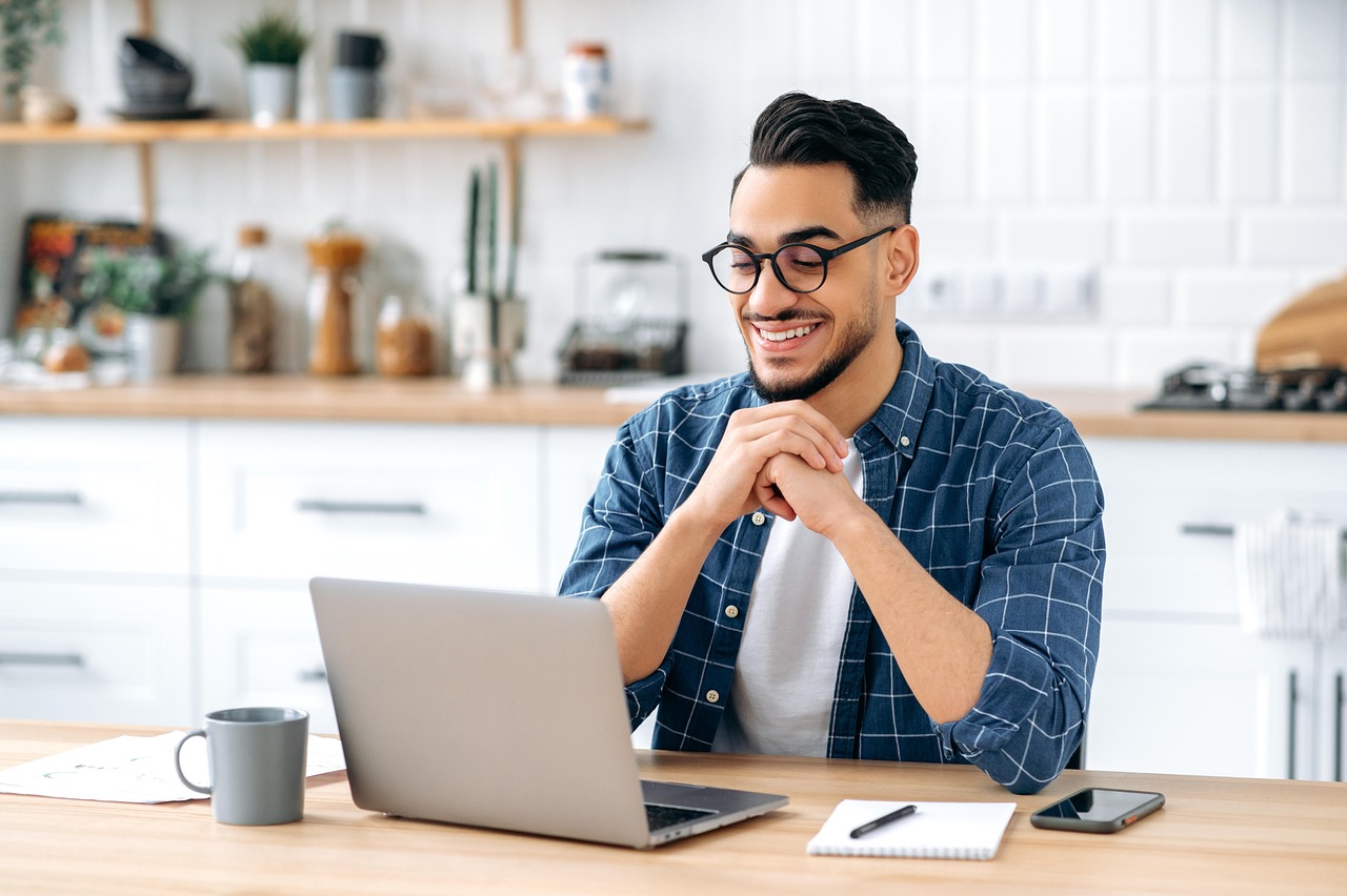 Etudiant souriant devant son ordinateur portable. En arrière plan, les éléments de l'intérieur de la maison, la cuisine.