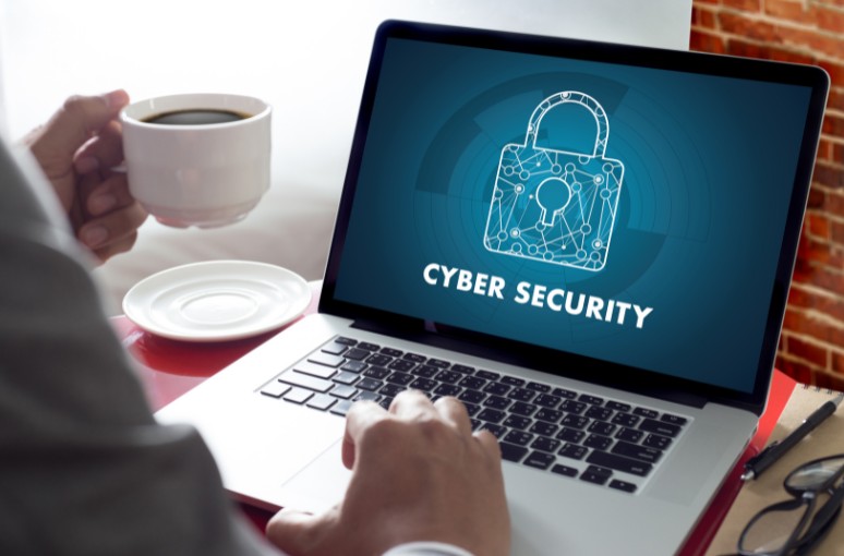 un ordinateur portable sur l'écran duquel figure un cadenas et la mention cyber security. On voit les mains de l'utilisateur. Il tient une tasse de café.