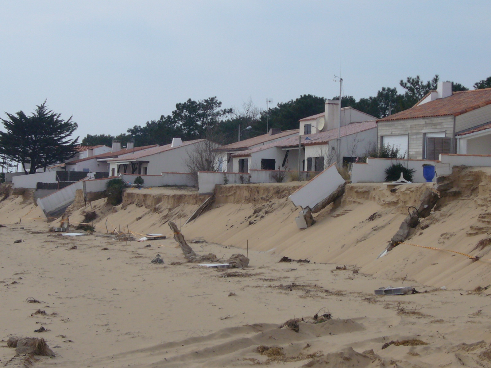 Maisons en bord de plage fragilisées par l’érosion côtière, avec dunes fortement entaillées et murs de protection effondrés.